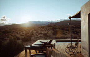 Um deck de madeira com cadeiras e uma cadeira de balanço cria uma área gourmet rústica, com vista para uma paisagem desértica ao pôr do sol, com montanhas ao longe e luz suave do sol projetando longas sombras.