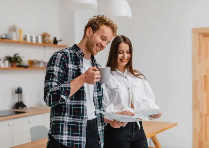 Imagem de um homem e uma mulher na cozinha de um imóvel em pé em frente à mesa lendo alguns documentos enquanto o homem toma café para ilustrar matéria sobre contrato de gaveta