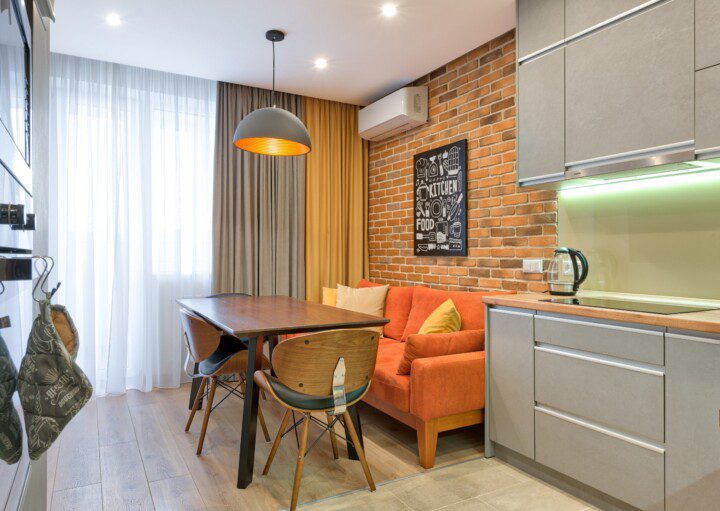 Foto de um exemplo de apartamento pequeno decorado. A imagem mostra um ambiente que integra cozinha com sala de estar e sala de jantar. Nela há um fogão embutido na parede, uma mesa com duas cadeiras, um sofá laranja e um armário de cozinha com pia embutida.