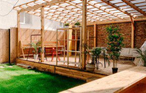 Um quintal moderno conta com um deck de madeira com pérgola, decoração elegante, mesas ao ar livre, vasos de plantas e um gramado verde. A área é cercada e delimitada por prédios de apartamentos.