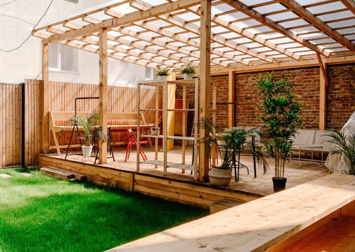 Um quintal moderno conta com um deck de madeira com pérgola, decoração elegante, mesas ao ar livre, vasos de plantas e um gramado verde. A área é cercada e delimitada por prédios de apartamentos.