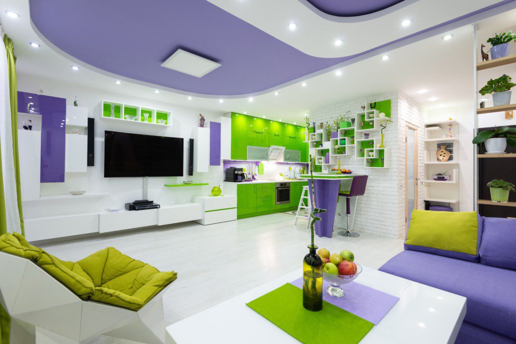 A foto mostra um exemplo de decoração retrô em um lar de conceito aberto com: poltrona, mesa de centro, rack, TV, bancada, armários de cozinha e muitos elementos decorativos.