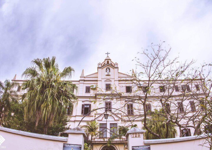 Foto que ilustra matéria sobre escolas em Novo Hamburgo (RS) mostra a fachada do Colégio Santa Catarina por trás das folhas verdes e galhos de árvores.