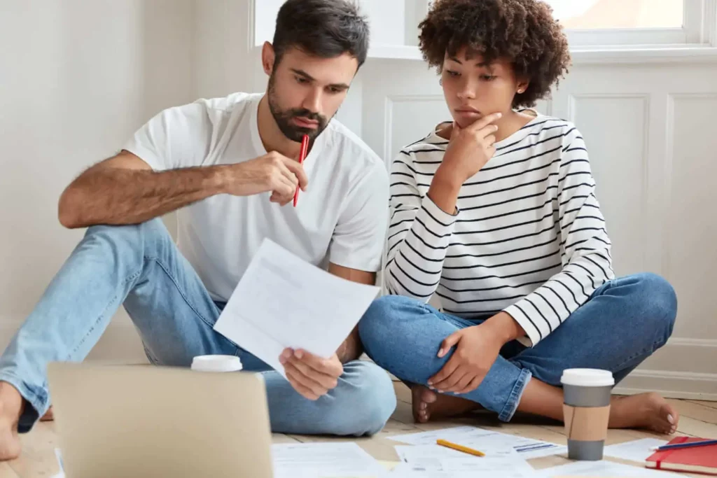 Imagem de um homem e uma mulher sentados no chão de um imóvel analisando as informações contidas em um documento para ilustrar matéria sobre o que é contrato de gaveta. No chão ainda estão outros papéis, um copo de café, um caderno, uma caneta e um notebook