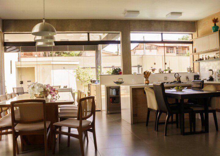 Área gourmet com mesa de jantar e espaço de cozinha ao fundo. Cadeiras, lustres e flores decoram o ambiente. Imagem disponível no Pexels