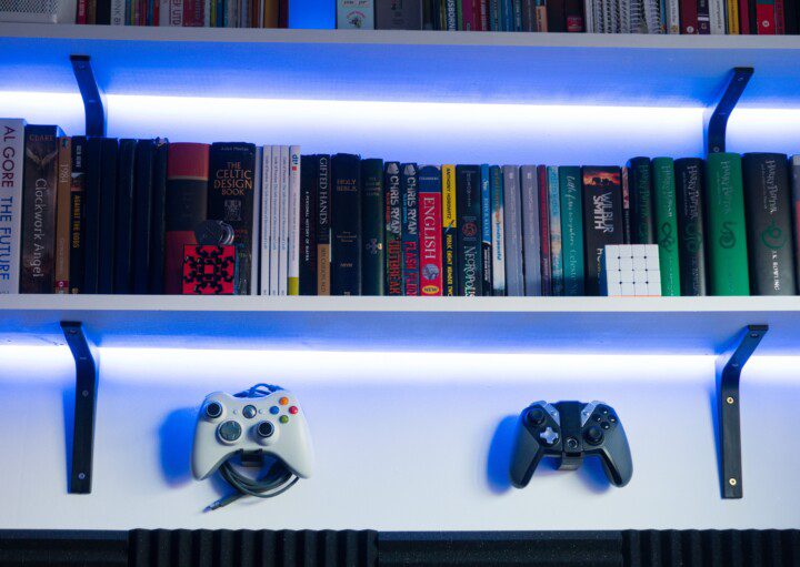 Uma prateleira com livros e um cubo mágico é iluminada por luzes LED azuis. Abaixo da prateleira, dois controles de videogame estão fixados na parede, com painéis de espuma acústica parcialmente visíveis na parte inferior.