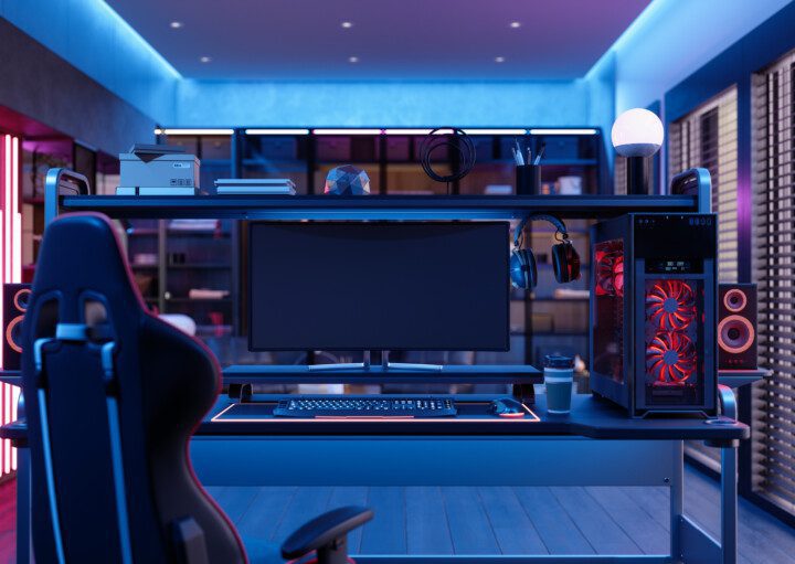 A foto mostra um exemplo de quarto gamer. Há uma poltrona gamer, uma mesa com monitor, teclado, mouse, fone de ouvido e CPU em cima dela. Há também uma iluminação especial no quarto, dando uma coloração em tom vermelho na lateral e azul neon no teto.