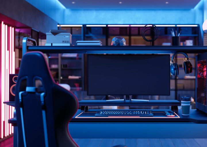 A foto mostra um exemplo de quarto gamer. Há uma poltrona gamer, uma mesa com monitor, teclado, mouse, fone de ouvido e CPU em cima dela. Há também uma iluminação especial no quarto, dando uma coloração em tom vermelho na lateral e azul neon no teto.