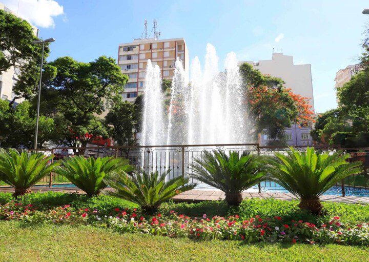 Foto que ilustra matéria sobre o que fazer em São José do Rio Preto mostra chafariz da Praça Rui Barbosa cercado por um gradeado, plantas, árvores e com prédios ao fundo em um dia de céu azul.