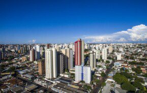 Foto que ilustra matéria sobre bairros de Uberlândia mostra uma visão do alto da cidade em um dia ensolarado de céu azul e poucas nuvens, com prédios muito altos no centro da imagem, algumas casas no canto inferior esquerdo e uma região arborizada no canto inferior direito.