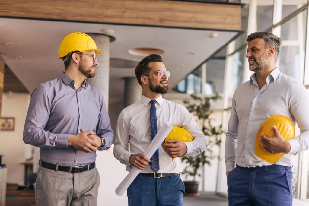 Imagem de três homens em trajes de negócios, dois segurando capacetes de construção amarelos e um segurando plantas, estão em um ambiente interno discutindo, provavelmente analisando um memorial descritivo de um projeto de construção