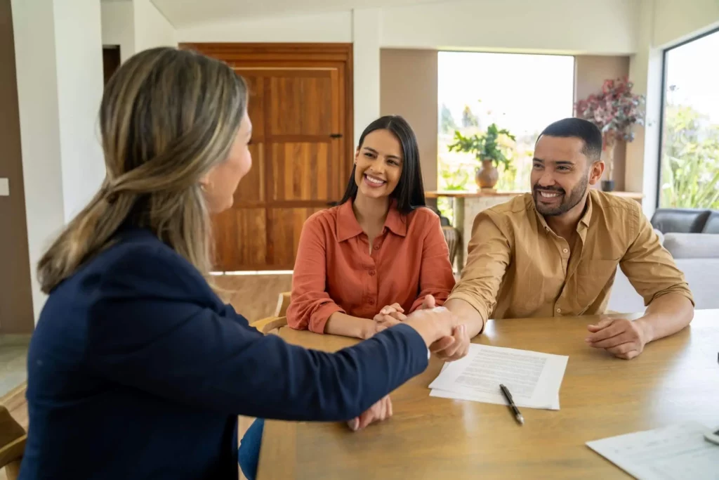 Imagem de um casal sorridente sentado em uma mesa com papéis, apertando as mãos de uma mulher em trajes de negócios em uma casa moderna e iluminada