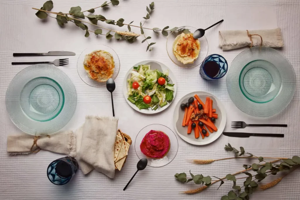 Imagem de uma mesa posta para duas pessoas mostra como montar uma mesa de jantar, com uma salada, um prato de cenoura e azeitonas, um molho rosa, pratos assados, pão achatado, taças de vinho, pratos e talheres sobre uma toalha de mesa branca, tudo realçado por folhagens verdes frescas.