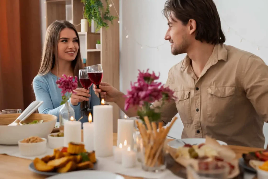 Imagem de uma mulher e um homem sentados à mesa arranjada e iluminada por velas, sorrindo e brindando com taças de vinho tinto. A mesa está posta com diversos pratos e decorada com flores e velas, servindo de inspiração para quem quiser aprender como montar uma mesa de jantar elegante para ocasiões especiais.
