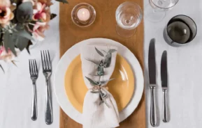 Imagem de uma mesa posta com um prato branco e amarelo, um guardanapo enrolado e amarrado com folhagens, três garfos, duas facas, dois copos, uma vela e um buquê de flores sobre uma toalha de mesa branca
