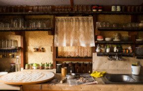 A foto mostra um exemplo de cozinha rústica em que há diferentes prateleiras de madeira ocupando toda a parede. Nelas tem utensílios como copos, xícaras, pratos, temperos, entre outros. Há também uma bancada de madeira com uma pia embutida.