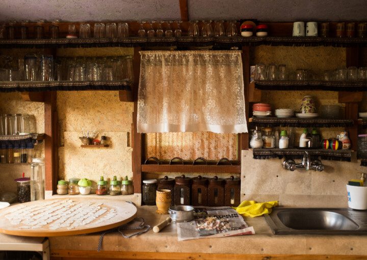 A foto mostra um exemplo de cozinha rústica em que há diferentes prateleiras de madeira ocupando toda a parede. Nelas tem utensílios como copos, xícaras, pratos, temperos, entre outros. Há também uma bancada de madeira com uma pia embutida.