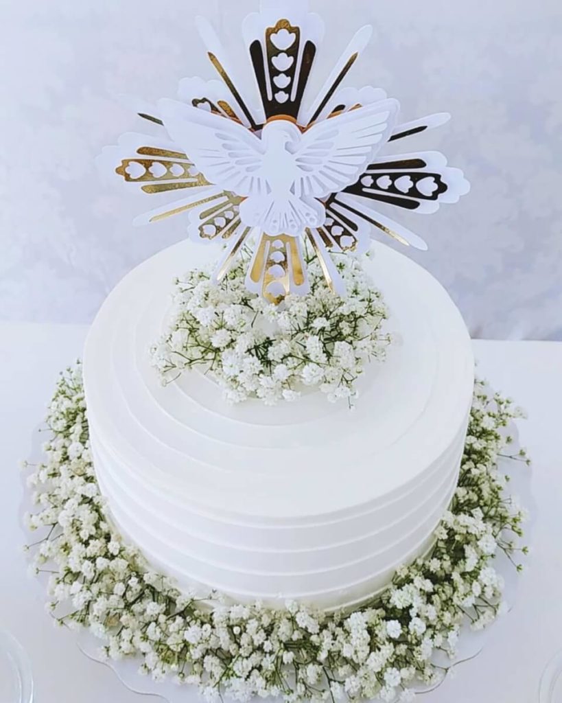 Um bolo redondo branco decorado com flores brancas de mosquitinho ao redor da base e no topo, com uma pomba de papel e uma cruz decorativa dourada e branca — perfeito para uma decoração batizado elegante.