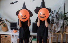 Foto que ilustra matéria sobre decoração de Halloween mostra duas crianças vestidas de preto, com chapéus de bruxa preto e com balões na cor laranja imitando abóboras com carinhas em frente a seus rostos. Na parede atrás deles, há pequenos morcegos feitos de papel pendurados.