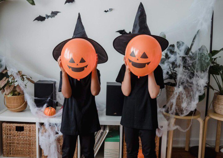 Foto que ilustra matéria sobre decoração de Halloween mostra duas crianças vestidas de preto, com chapéus de bruxa preto e com balões na cor laranja imitando abóboras com carinhas em frente a seus rostos. Na parede atrás deles, há pequenos morcegos feitos de papel pendurados.