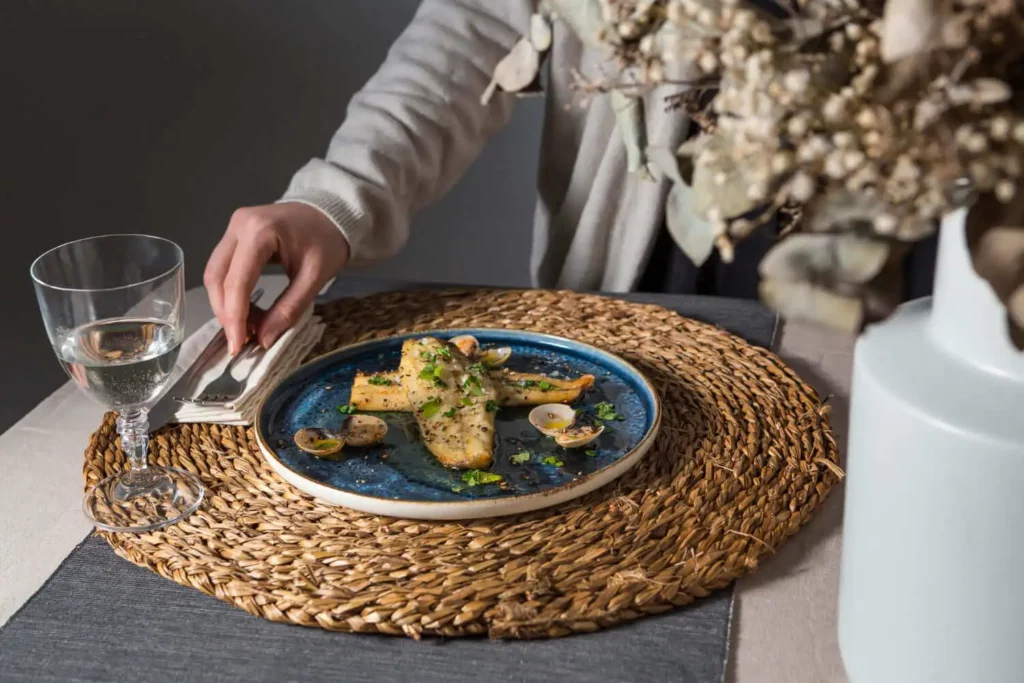 Imagem de uma pessoa pegando um guardanapo ao lado de um prato de peixe e mariscos guarnecidos com ervas, servido em um prato azul sobre um jogo americano de tecido, ao lado de um copo de água e um vaso de flores secas