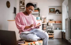 Imagem de uma pessoa usando óculos e um suéter rosa está sentada em uma cozinha, usando um smartphone enquanto segura uma caneta com cadernos, papéis e um prato de comida na mesa à sua frente
