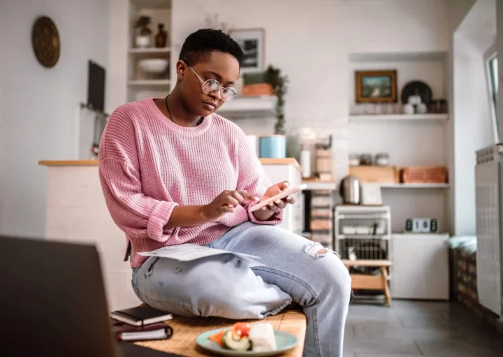 Imagem de uma pessoa usando óculos e um suéter rosa está sentada em uma cozinha, usando um smartphone enquanto segura uma caneta com cadernos, papéis e um prato de comida na mesa à sua frente
