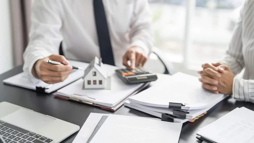 Imagem de duas pessoas em trajes de negócios sentadas em uma mesa com papéis, uma calculadora e uma casa modelo, discutindo documentos