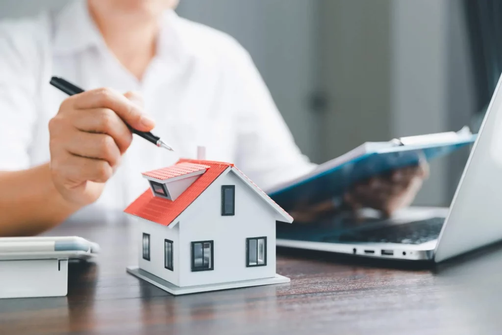 Imagem de uma pessoa segurando uma caneta revisando documentos em frente a um laptop, com uma pequena maquete de casa colocada sobre a mesa