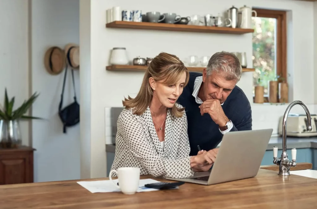 Imagem de uma mulher e um homem sentados juntos em um balcão de cozinha, olhando atentamente para a tela de um laptop. Papéis, uma caneta e uma caneca de café estão sobre o balcão. Prateleiras com pratos são visíveis ao fundo