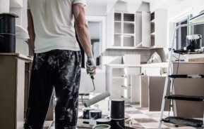 A imagem mostra um homem reformando uma cozinha. Ele tem a roupa manchada de tinta e segura um rolo de tinta. Há na imagem uma cozinha com armários embutidos e também elementos usados na reforma espalhados: galões de tinta, escada, sacolas e outros.