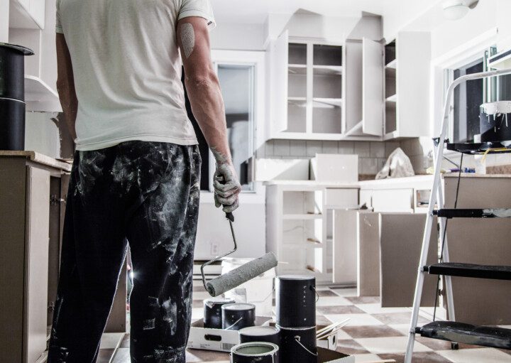 A imagem mostra um homem reformando uma cozinha. Ele tem a roupa manchada de tinta e segura um rolo de tinta. Há na imagem uma cozinha com armários embutidos e também elementos usados na reforma espalhados: galões de tinta, escada, sacolas e outros.