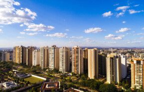 Foto que ilustra matéria sobre bairros de Ribeirão Preto mostra uma visão do alto da cidade, com diversos prédios grandes no centro da imagem, áreas arborizadas no canto inferior esquerdo e um céu azul com poucas nuvens brancas de um dia claro.