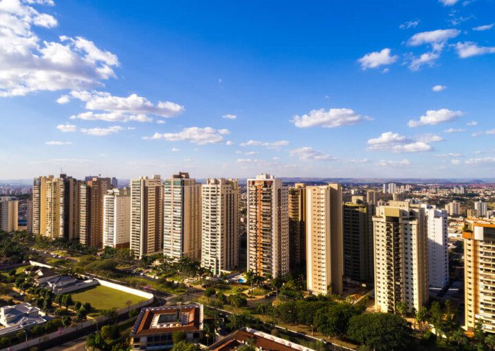 Foto que ilustra matéria sobre bairros de Ribeirão Preto mostra uma visão do alto da cidade, com diversos prédios grandes no centro da imagem, áreas arborizadas no canto inferior esquerdo e um céu azul com poucas nuvens brancas de um dia claro.