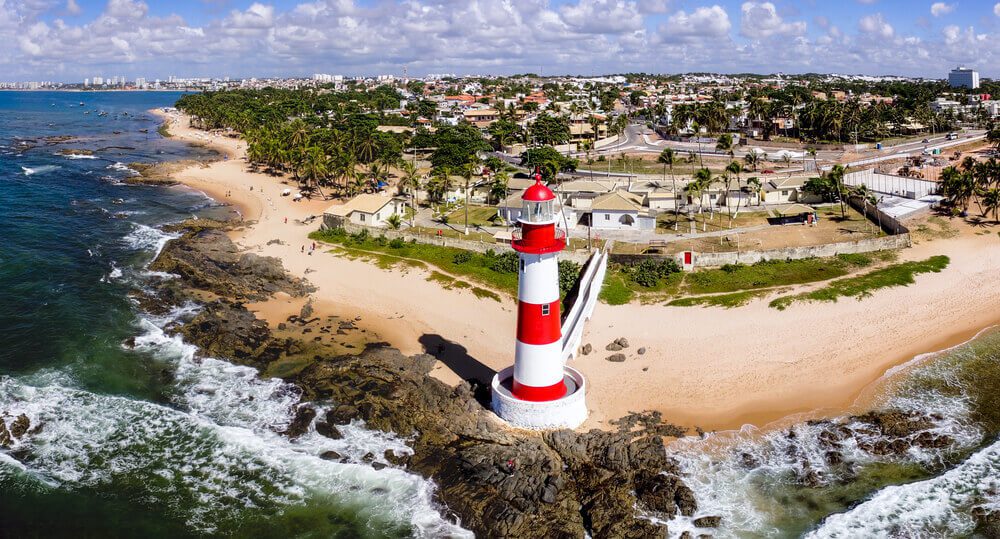 Foto que ilustra matéria sobre bairros mais seguros de Salvador mostra um farol à beira mar pintado de grossas listras vermelhas e brancas. Atrás dele está o bairro de Itapuã.