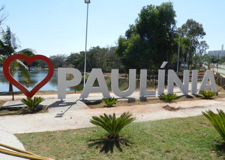 Foto que ilustra matéria sobre como chegar em Paulínia mostra uma imagem de um letreiro em um parque com um coração vermelho e o nome da cidade escrito em branco. Atrás, aparecem um lago, árvores e um céu azul claro.