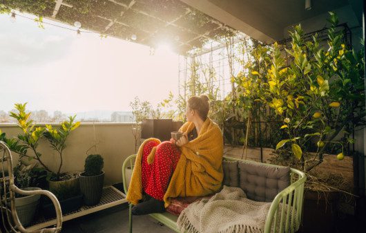 Foto de uma varanda com uma mulher sentada no sofá. Ao redor dela há plantas de diferentes tamanhos.