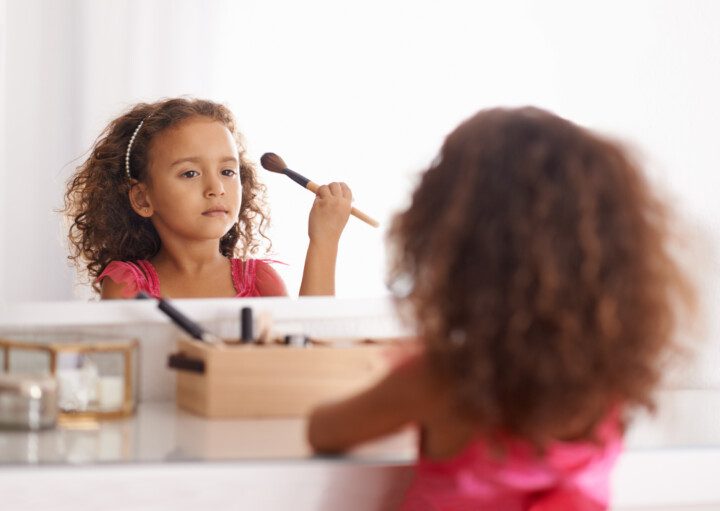 Imagem de uma menina segurando um pincel de maquiagem sentada de frente para uma penteadeira.