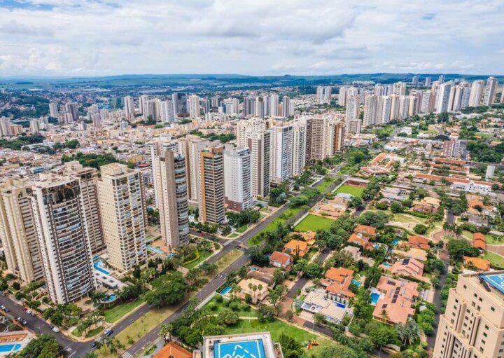 Foto que ilustra matéria sobre morar em Ribeirão Preto mostra uma visão do alto da Avenida Professor João Fiúza, a mais importante da cidade, com muitos prédios altos à esquerda e casas em zonas arborizadas à direita.