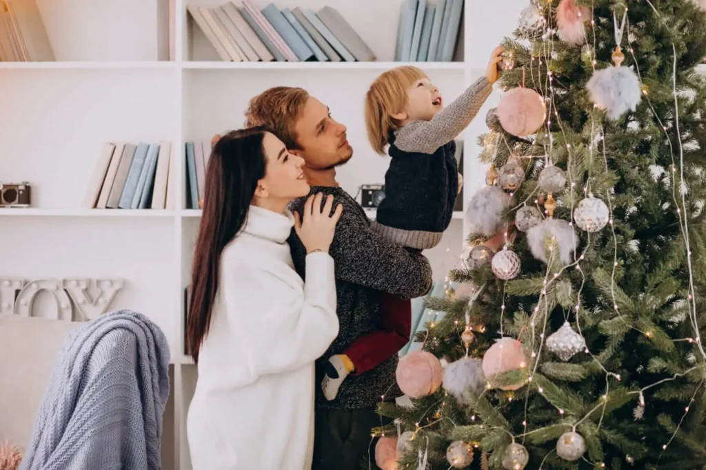 Imagem de uma família decorando uma árvore de Natal juntos. Um homem segura uma criança pequena que tenta pendurar um enfeite, enquanto uma mulher está próxima, sorrindo em sua sala de estar aconchegante e festivamente iluminada.