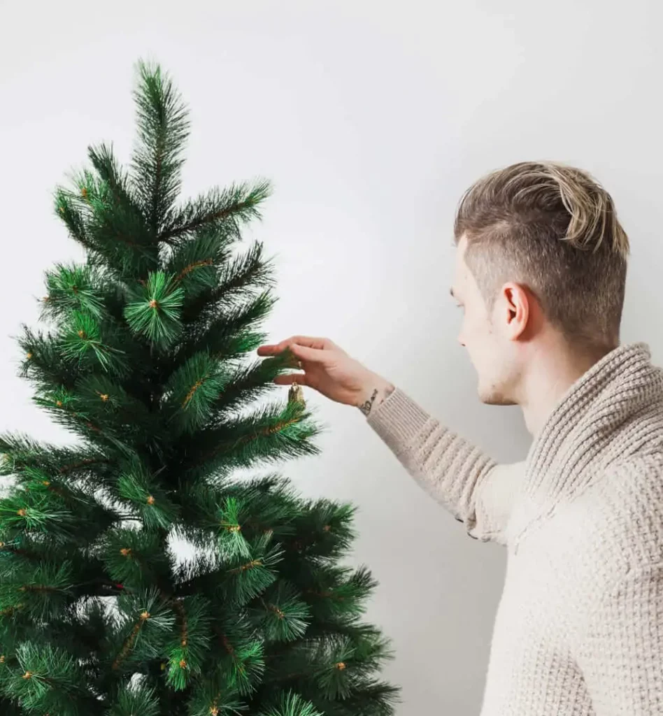 Imagem de uma pessoa com cabelos curtos e claros e um suéter bege arrumando os galhos de uma árvore de Natal