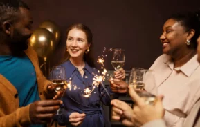 Imagem de um grupo de pessoas sorrindo e comemorando em um ambiente fechado, segurando taças de vinho branco e espumantes, com balões dourados e decoração de ano novo dando um toque festivo ao fundo.