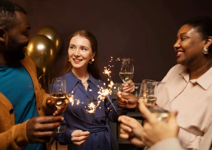 Imagem de um grupo de pessoas sorrindo e comemorando em um ambiente fechado, segurando taças de vinho branco e espumantes, com balões dourados e decoração de ano novo dando um toque festivo ao fundo.