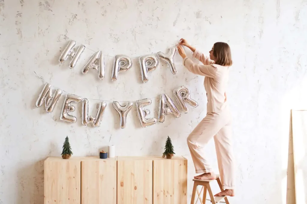 Imagem de uma mulher de pijama claro em um banquinho de madeira, pendurando balões prateados com letras HAPPY NEW YEAR em uma parede branca acima de um armário de madeira com pequenas decorações.