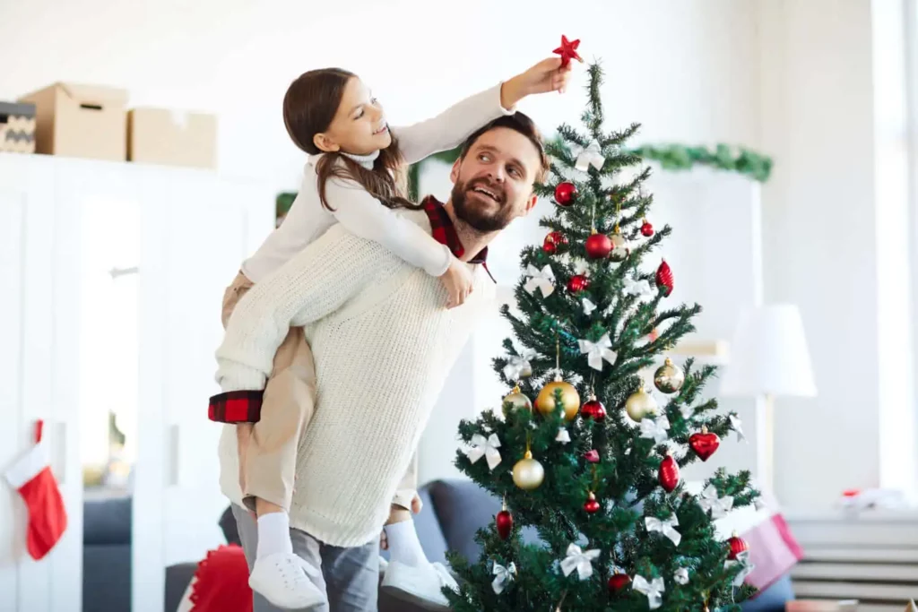 Imagem de um homem e uma menina enfeitando uma árvore de Natal em uma sala de estar brilhante e festiva.