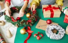 Imagem de duas pessoas decorando uma guirlanda de Natal em uma mesa coberta com presentes embrulhados, um bloco de notas e uma caneta, um carretel de barbante, uma tesoura, enfeites de Natal e materiais que mostram como fazer presentes de Natal.