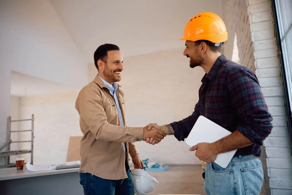 Imagem de dois homens, um com um capacete e segurando um tablet, o outro sem capacete, apertam as mãos e sorriem dentro de um prédio bem iluminado, parcialmente construído