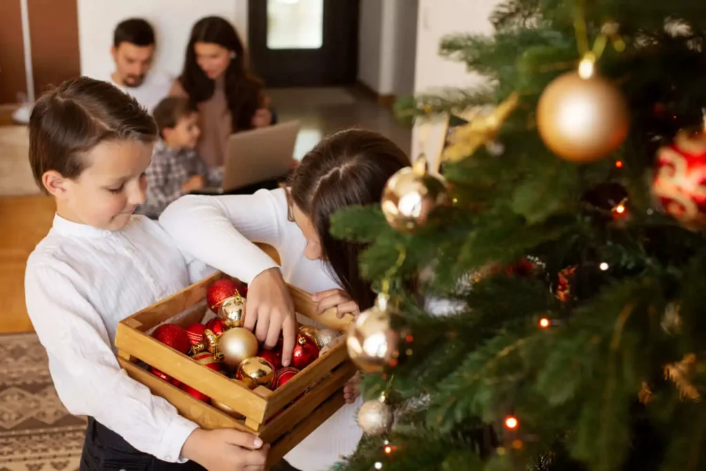 Imagem de duas crianças segurando uma caixa de madeira cheia de enfeites de Natal perto de uma árvore decorada, enquanto dois adultos se sentam ao fundo usando um laptop.