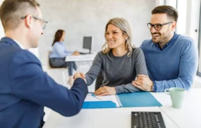 Imagem de uma mulher acompanhada de um homem em um escritório de finanças apertando a mão de um homem enquanto segura uma pasta com documentos para ilustrar matéria sobre interveniente quitante