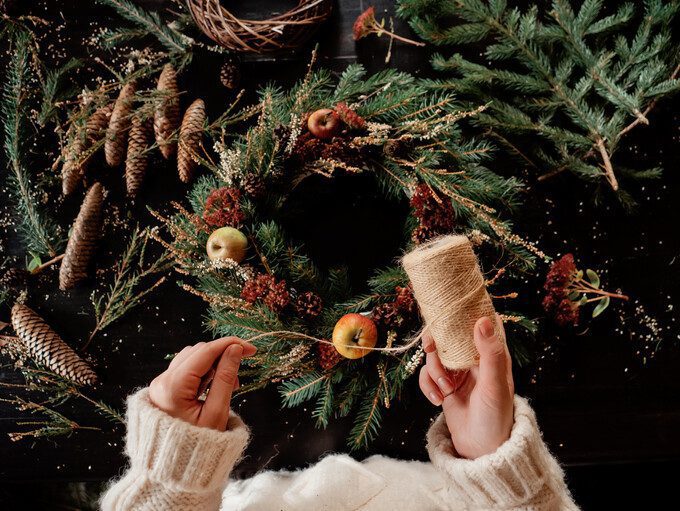 Maos brancas segurando um rolo de sisal com uma guirlanda de natal ao fundo com macas.